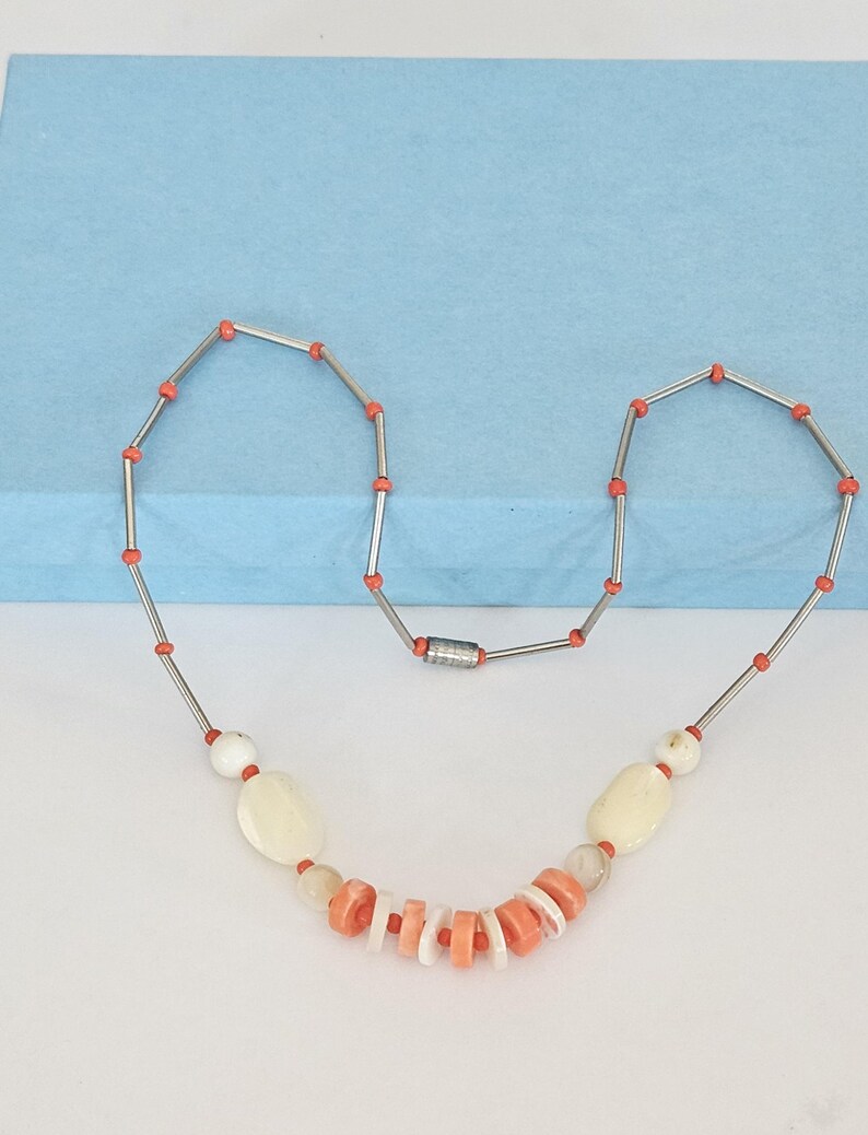 May include: A necklace featuring a series of silver-toned metal tubes separated by small orange beads. The center section includes larger off-white oval beads, translucent round beads, and a cluster of alternating flat orange and white disc beads.
