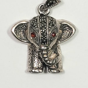 May include: A silver elephant pendant with marcasite detailing. The elephant has red gemstone eyes and a textured surface. The pendant is designed with a loop for a chain.