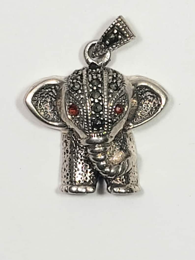 May include: A silver elephant pendant with marcasite detailing. The elephant has red gemstone eyes and a textured surface. The pendant is designed with a loop for a chain.