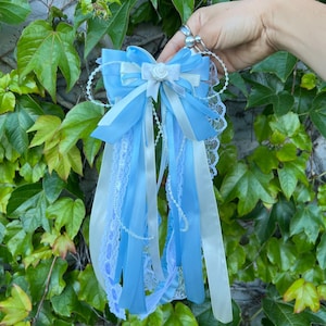 May include: A light blue and white decorative bow with long ribbons and lace trim. The bow features a small white rose in the center and is held against a backdrop of green leaves.