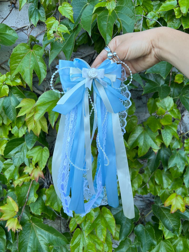 May include: A light blue and white decorative bow with long ribbons and lace trim. The bow features a small white rose in the center and is held against a backdrop of green leaves.