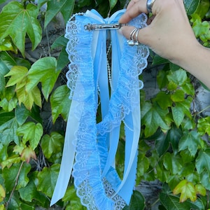 May include: A light blue hair bow with white and light blue ribbons and lace trim. The hair accessory is held by a silver barrette clip. The background is green ivy.