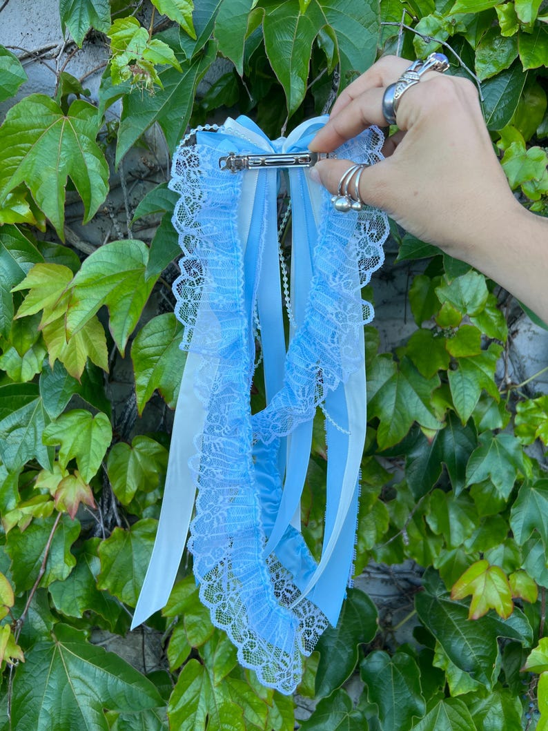 May include: A light blue hair bow with white and light blue ribbons and lace trim. The hair accessory is held by a silver barrette clip. The background is green ivy.