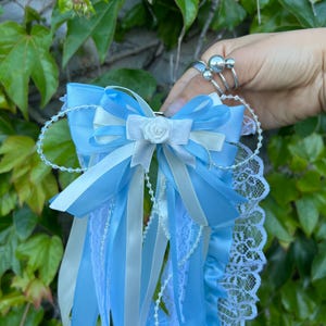 May include: A light blue and white satin garter with a bow and lace trim. The garter features a small white rose and is adorned with ribbons and beaded accents. The background is a lush green foliage.