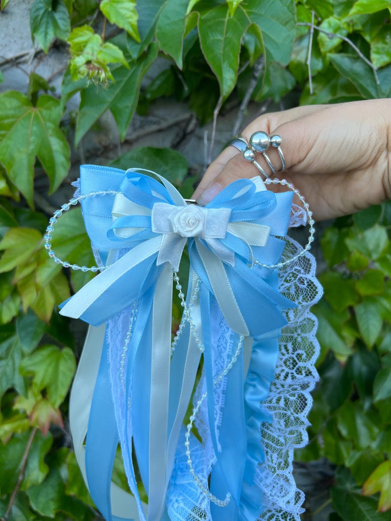 May include: A light blue and white satin garter with a bow and lace trim. The garter features a small white rose and is adorned with ribbons and beaded accents. The background is a lush green foliage.
