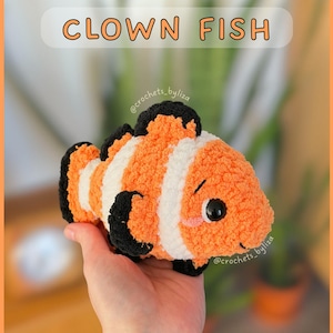 Puede incluir: Un pez payaso de ganchillo en naranja, blanco y negro. El pez tiene una cara sonriente y se sostiene en una mano. El texto "CLOWN FISH" está encima del pez, con el nombre de la tienda debajo.