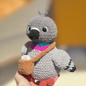 Op de afbeelding: Een grijs gehaakte duif knuffel met een zwarte snavel, donkere ogen en een kleine kuif op de kop. De vogel heeft een levendig blauwgroen en magentakleurig patroon in de nek, oranje pootjes en een bruine schoudertas met een wit item.