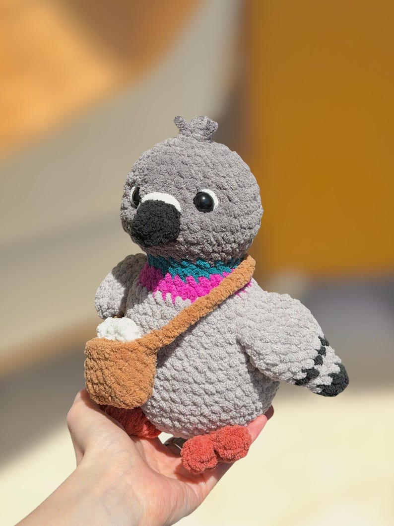 Op de afbeelding: Een grijs gehaakte duif knuffel met een zwarte snavel, donkere ogen en een kleine kuif op de kop. De vogel heeft een levendig blauwgroen en magentakleurig patroon in de nek, oranje pootjes en een bruine schoudertas met een wit item.