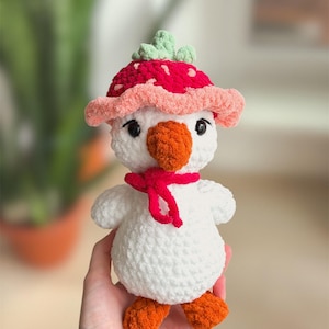 May include: A handmade white plush duck toy with orange feet and beak, wearing a red scarf and a strawberry-shaped hat with green leaves. The toy is soft and cuddly, perfect for a child's playtime or nursery decor.