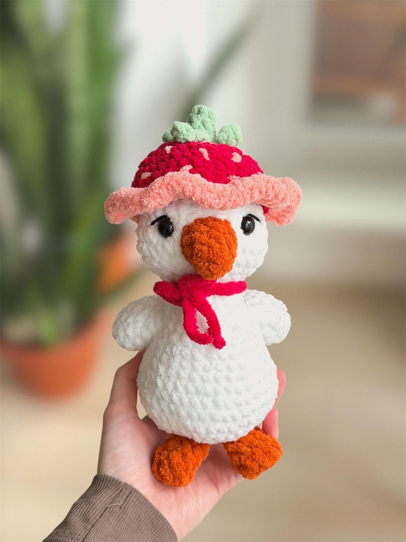 May include: A handmade white plush duck toy with orange feet and beak, wearing a red scarf and a strawberry-shaped hat with green leaves. The toy is soft and cuddly, perfect for a child's playtime or nursery decor.