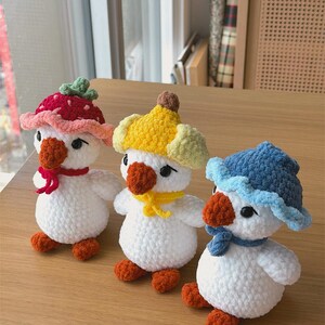 May include: Three handmade crochet duck toys with white bodies, orange feet and beaks, and black eyes. Each duck wears a different hat: a red strawberry hat, a yellow banana hat, and a blue hat. They are arranged on a wooden surface.
