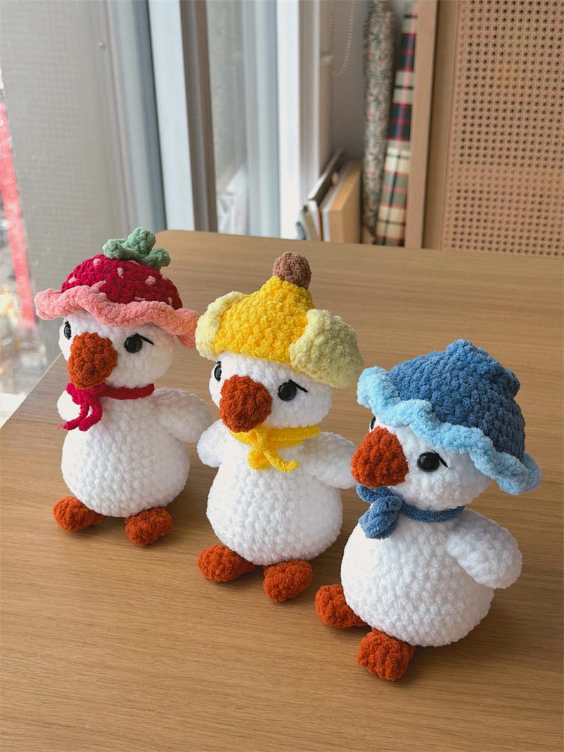 May include: Three handmade crochet duck toys with white bodies, orange feet and beaks, and black eyes. Each duck wears a different hat: a red strawberry hat, a yellow banana hat, and a blue hat. They are arranged on a wooden surface.