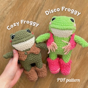 May include: Two crocheted frog plushies. The left frog is olive green with a brown vest and the text Cozy Froggy. The right frog is bright green with a pink scarf and boots and the text Disco Froggy. Both have white bellies and black eyes.