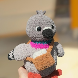 Op de afbeelding: Een grijs gehaakte duif knuffel met een zwarte snavel, ronde zwarte ogen en oranje voeten. De vogel heeft een felroze en turquoise halsband en draagt een kleine bruine gehaakte schoudertas met daarin een wit rechthoekig voorwerp.