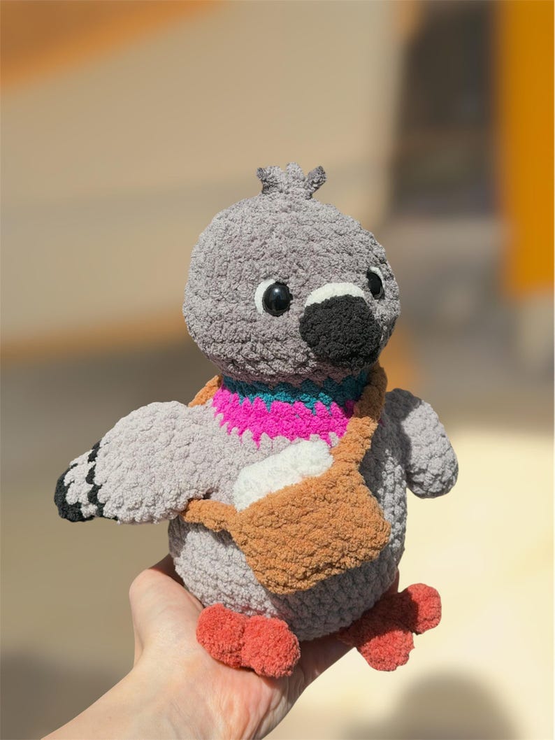 Op de afbeelding: Een grijs gehaakte duif knuffel met een zwarte snavel, ronde zwarte ogen en oranje voeten. De vogel heeft een felroze en turquoise halsband en draagt een kleine bruine gehaakte schoudertas met daarin een wit rechthoekig voorwerp.