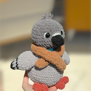 Op de afbeelding: Een gehaakt duif knuffeltje met een zacht grijs lichaam, een donkergrijze snavel en feloranje pootjes. De duif draagt een getextureerde beige sjaal om de nek, heeft kleine zwarte plastic oogjes en een klein plukje grijs garen op de kop.