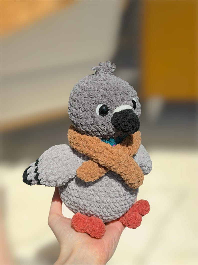 Op de afbeelding: Een gehaakt duif knuffeltje met een zacht grijs lichaam, een donkergrijze snavel en feloranje pootjes. De duif draagt een getextureerde beige sjaal om de nek, heeft kleine zwarte plastic oogjes en een klein plukje grijs garen op de kop.