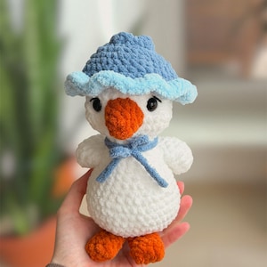 May include: A handmade crochet snowman toy. The snowman is white with an orange beak and feet. It wears a blue hat and scarf. The toy is held in a person's hand.