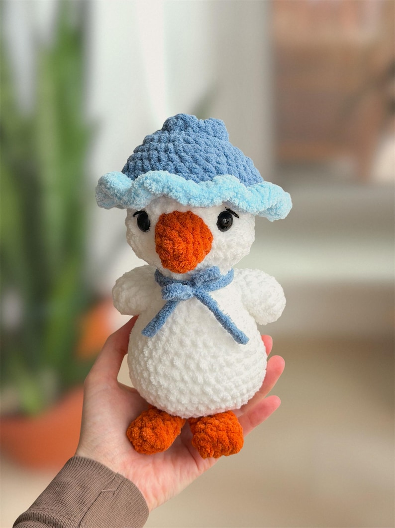 May include: A handmade crochet snowman toy. The snowman is white with an orange beak and feet. It wears a blue hat and scarf. The toy is held in a person's hand.