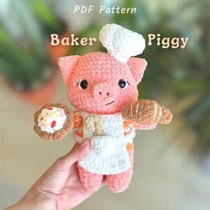 Puede incluir: Un cerdo rosa hecho a mano con ganchillo, con un gorro y delantal de chef blancos, sosteniendo un cruasán y un pastel. El cerdo tiene ojos negros y una expresión sonriente. El texto "PDF Pattern" y "Baker Piggy" son visibles.