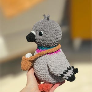 Op de afbeelding: Een gehaakt duif knuffeltje met een grijs, getextureerd lichaam, een zwarte snavel en oranje pootjes. De vogel heeft een halsband in turquoise en roze en draagt een klein beige tasje met een wit voorwerp erin. De vleugels hebben zwart-witte details.