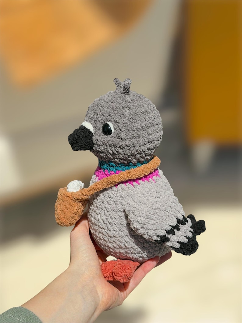 Op de afbeelding: Een gehaakt duif knuffeltje met een grijs, getextureerd lichaam, een zwarte snavel en oranje pootjes. De vogel heeft een halsband in turquoise en roze en draagt een klein beige tasje met een wit voorwerp erin. De vleugels hebben zwart-witte details.