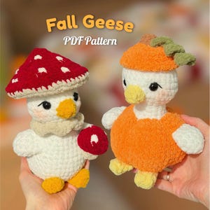 Peut inclure: Deux oies en peluche crochetées. L'une porte un chapeau champignon rouge et blanc et tient un petit champignon rouge. L'autre porte un chapeau citrouille orange avec une tige verte. Le texte indique Fall Geese PDF Pattern.