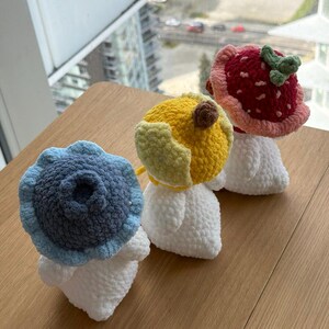 May include: Three handmade crochet mushroom figures. One is blue, resembling a blueberry, another is yellow, and the third is red, resembling a strawberry. Each mushroom has a white stem and is displayed on a wooden surface.