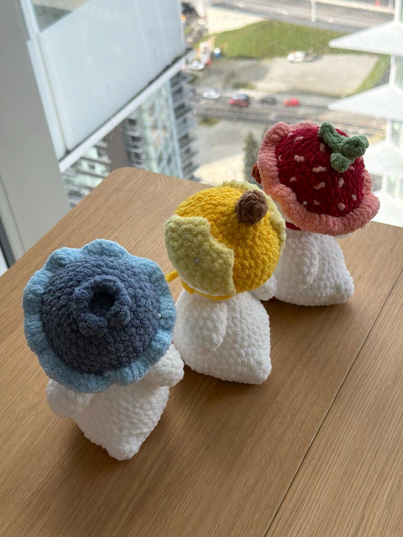 May include: Three handmade crochet mushroom figures. One is blue, resembling a blueberry, another is yellow, and the third is red, resembling a strawberry. Each mushroom has a white stem and is displayed on a wooden surface.