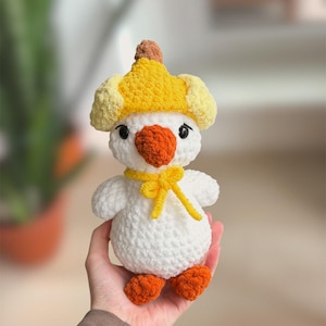 May include: A handmade white crochet duck toy with orange feet and beak, wearing a yellow hat and bow tie. The duck has black eyes and a fluffy texture, perfect for a child's toy or nursery decor.