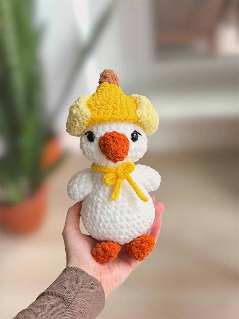 May include: A handmade white crochet duck toy with orange feet and beak, wearing a yellow hat and bow tie. The duck has black eyes and a fluffy texture, perfect for a child's toy or nursery decor.