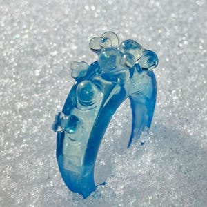 May include: A translucent blue ring with a bubbly design. The ring is made of a clear, light blue material and features several rounded, bubble-like embellishments on top. The ring is set against a snowy background.