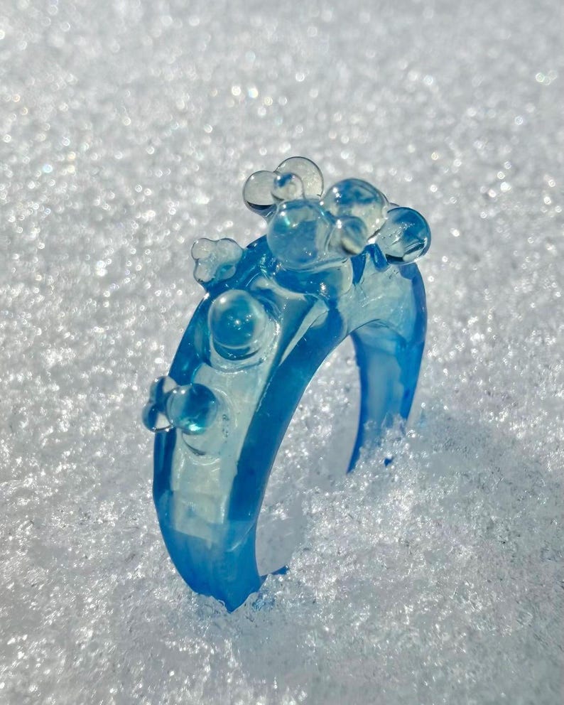 May include: A translucent blue ring with a bubbly design. The ring is made of a clear, light blue material and features several rounded, bubble-like embellishments on top. The ring is set against a snowy background.