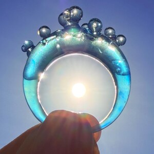 May include: A translucent blue ring with a bubbly design on top. The ring is held up to the sun, which shines through the center. The ring's color shifts from blue to green, with a clear sky in the background.