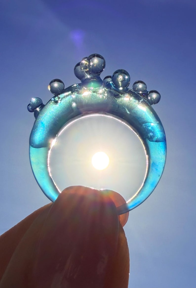 May include: A translucent blue ring with a bubbly design on top. The ring is held up to the sun, which shines through the center. The ring's color shifts from blue to green, with a clear sky in the background.