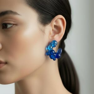 Könnte beinhalten: Ein Paar blaue Creolen aus Harz. Die Ohrringe weisen einen Farbverlauf von Blautönen auf, wobei eine Ansammlung von abgerundeten Formen ein einzigartiges Design bildet. Die Ohrringe werden am Ohr einer Person getragen, wobei der Fokus auf dem Schmuck liegt.