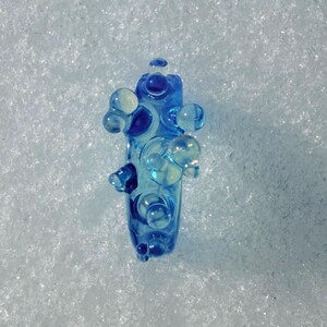 May include: A blue glass art piece with a unique, abstract design. The piece features rounded, bubble-like protrusions and a central column, all in shades of blue and clear glass. The object is set against a white, textured background.