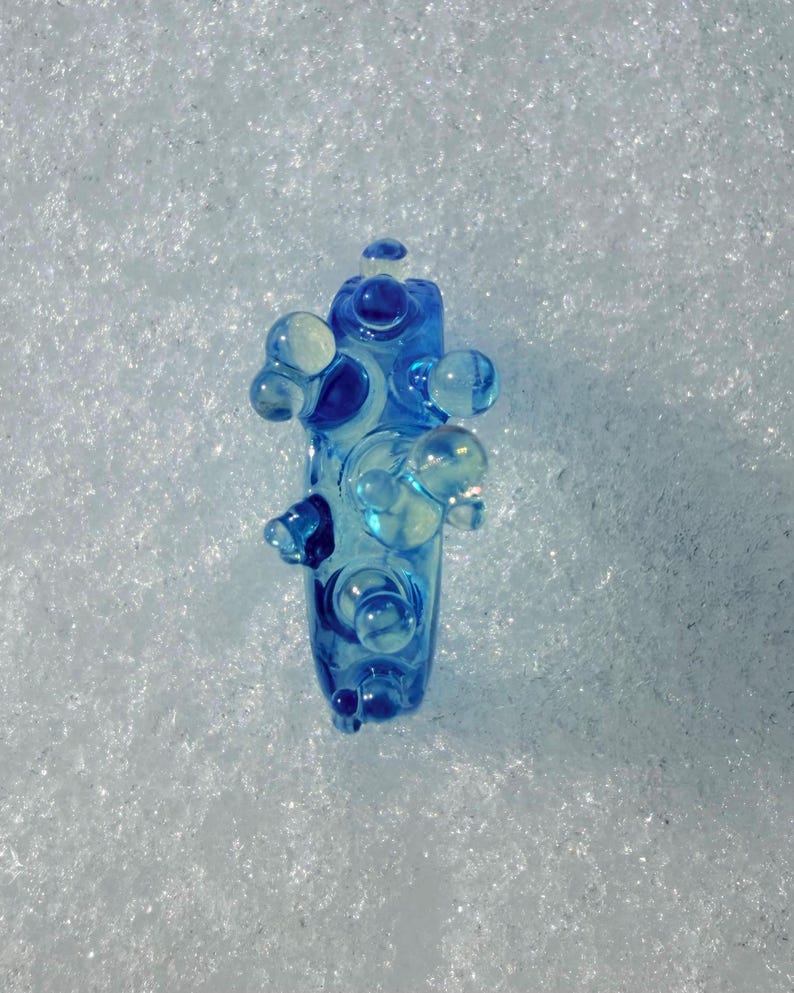 May include: A blue glass art piece with a unique, abstract design. The piece features rounded, bubble-like protrusions and a central column, all in shades of blue and clear glass. The object is set against a white, textured background.