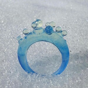 May include: A translucent blue ring with a bubbly design on top. The ring is a gradient of light to dark blue. The ring is set against a white, textured background.