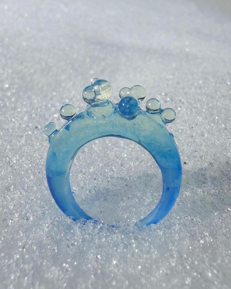 May include: A translucent blue ring with a bubbly design on top. The ring is a gradient of light to dark blue. The ring is set against a white, textured background.