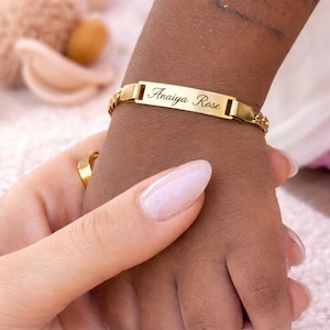 May include: A gold-toned baby bracelet featuring a central rectangular identification plate engraved with the name Anaiya Rose in a cursive script. The bracelet is attached to a delicate chain link band and includes text stating Personalized Baby Bracelet.