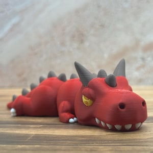 May include: A red dragon figurine with gray horns and spikes. The dragon has a detailed, textured surface, yellow eyes, and white teeth. The figurine is lying down on a wooden surface.