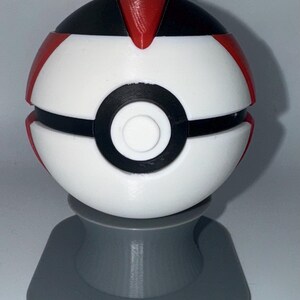 May include: A white, black, and red spherical object, resembling a ball, is displayed on a gray stand. The ball has a black band around the middle and red accents. The stand is a solid gray color.