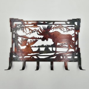 May include: Metal wall art featuring a moose scene with mountains and trees. The artwork has a rustic, copper-colored finish and includes four hooks for hanging items. The design depicts two moose in a natural landscape.