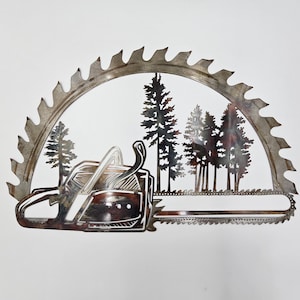 May include: Metal wall art featuring a chainsaw and a saw blade with a silhouette of trees in the background. The chainsaw is brown and silver, and the saw blade is silver with a black outline. The trees are black and are arranged in a row.