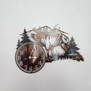 May include: Metal wall clock featuring a detailed elk silhouette with antlers, set against a mountain and forest scene. The clock face has a bronze finish with black numerals and gold hands. The clock is a decorative piece.