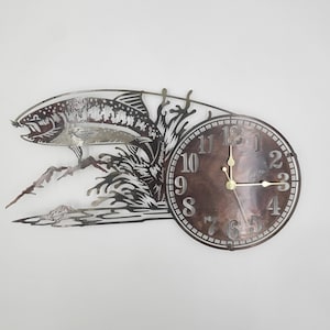 May include: Metal wall clock with a silhouette of a fish jumping out of the water. The clock face is brown with white numbers and hands.