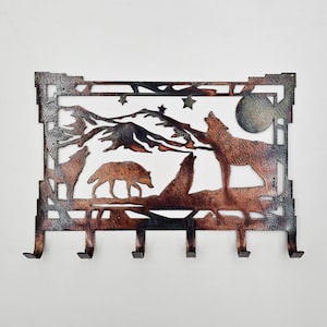 May include: Metal key holder with a rustic design featuring a mountain scene with three wolves howling at the moon. The key holder has five hooks.