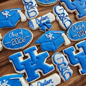 May include: An assortment of decorated sugar cookies in blue and white. The cookies include graduation caps, diploma scrolls, and cookies with the text "Class of 2026" and "Dylan". The cookies are arranged on a wooden surface.