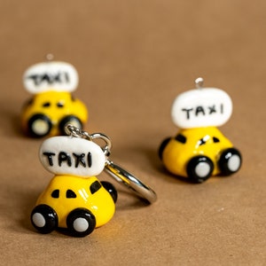 May include: Yellow taxi cab keychains with black wheels and a white top that reads "TAXI" in black letters. The keychains are made of a glossy material and have a silver-colored keyring attached. The background is a solid brown color.